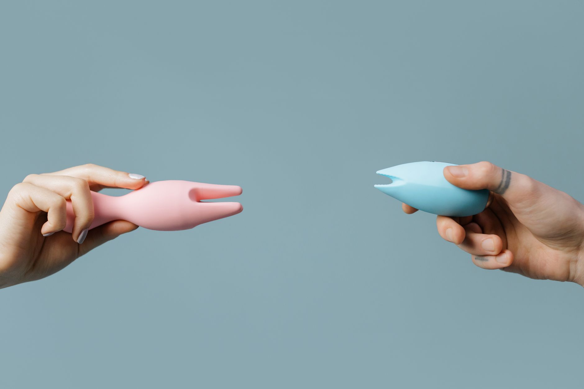 Hands holding pink and blue silicone vibrators against a soft pastel background